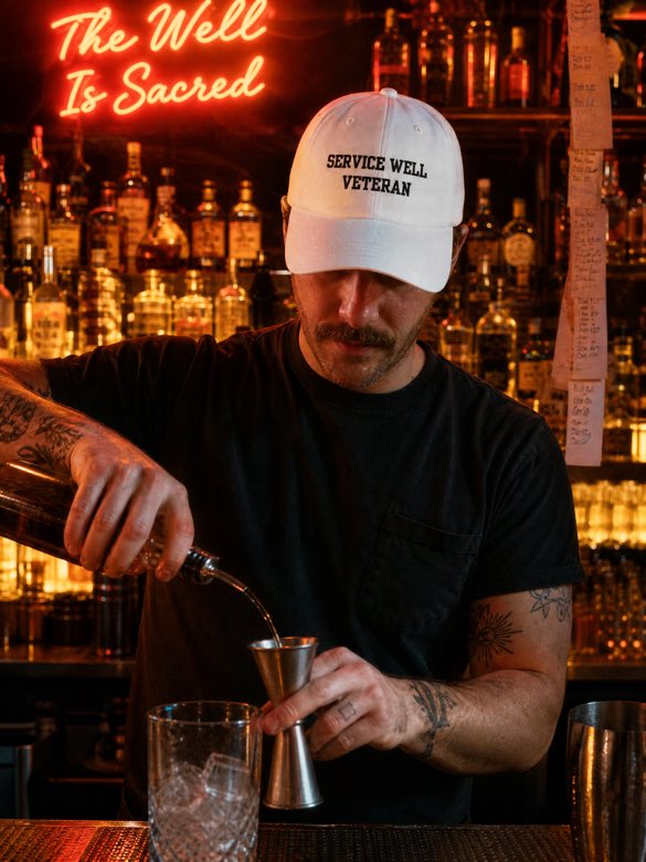 Bartender pouring a drink under a neon sign reading The Well Is Sacred